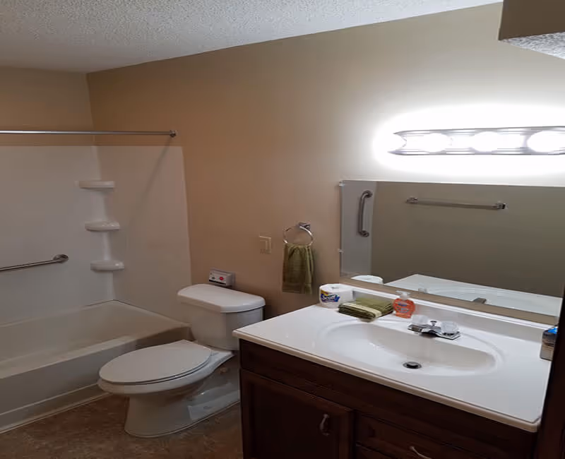 A clean bathroom with a white bathtub and shower combination on the left, a white toilet next to it, and a white sink with a wooden cabinet below on the right. Above the sink is a large mirror with a light fixture above it. A green hand towel hangs on a ring on the wall, and various toiletries are placed on the sink counter.