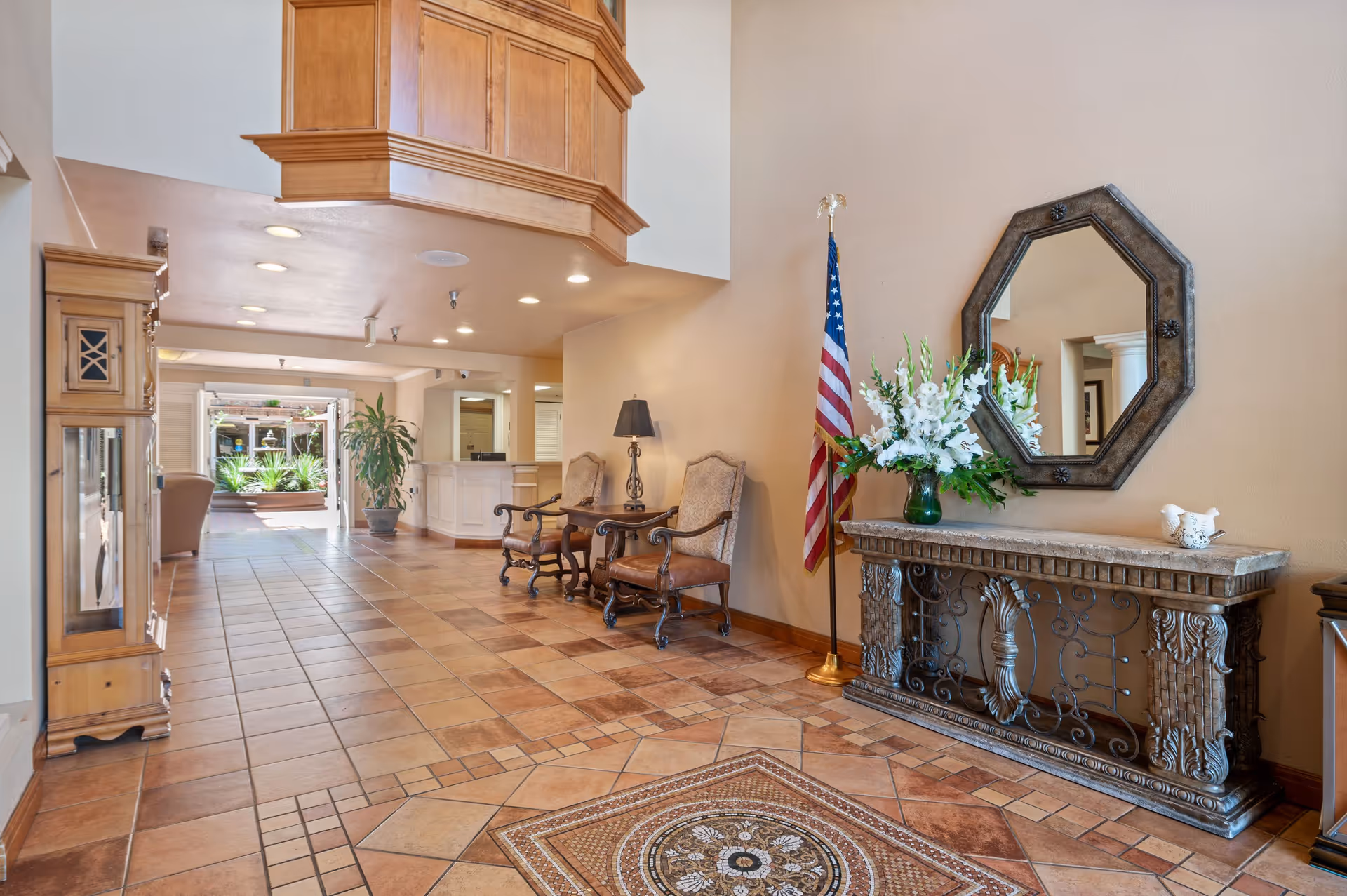 A spacious and well-lit interior hallway of a senior living community featuring a decorative tiled floor with a mosaic pattern, two ornate chairs with a small table and lamp between them, an American flag, a large mirror above a decorative console table with a vase of white flowers, and a wooden grandfather clock. The hallway leads to an open area with plants and natural light.