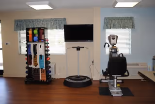 A small exercise room with wooden flooring featuring a rack of colorful dumbbells and exercise bands, a vibration plate machine, a recumbent exercise bike, and two windows with floral valances allowing natural light.