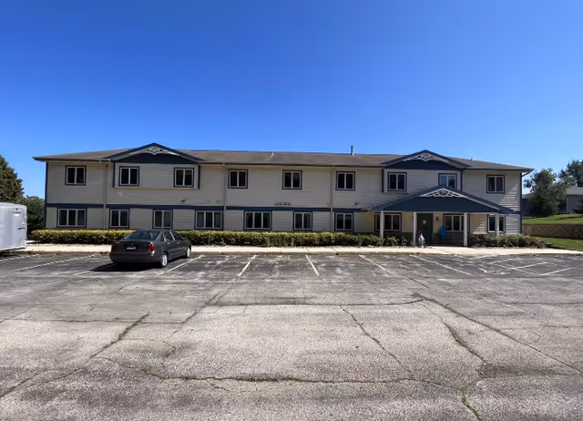 Exterior view of a two-story senior living facility building with beige siding and blue trim under a clear blue sky. There is a parking lot in front with one parked car and a small trailer on the left side. The entrance has a covered porch with decorative trim.