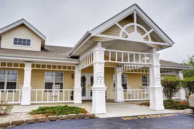 Exterior view of a single-story building with a covered entrance supported by white columns. The building has beige siding, multiple windows, and a small garden area with shrubs near the entrance. There are signs on the columns indicating 'No Parking Fire Lane' and the driveway has 'NO PARKING' painted on the asphalt.