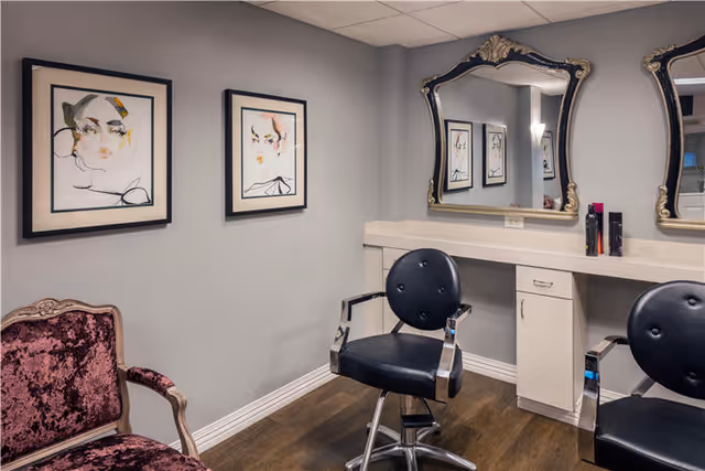 Interior view of a salon area with two black salon chairs in front of a white counter with drawers and two ornate framed mirrors. There is a vintage-style purple velvet armchair on the left side and two framed abstract face artworks on the gray wall.