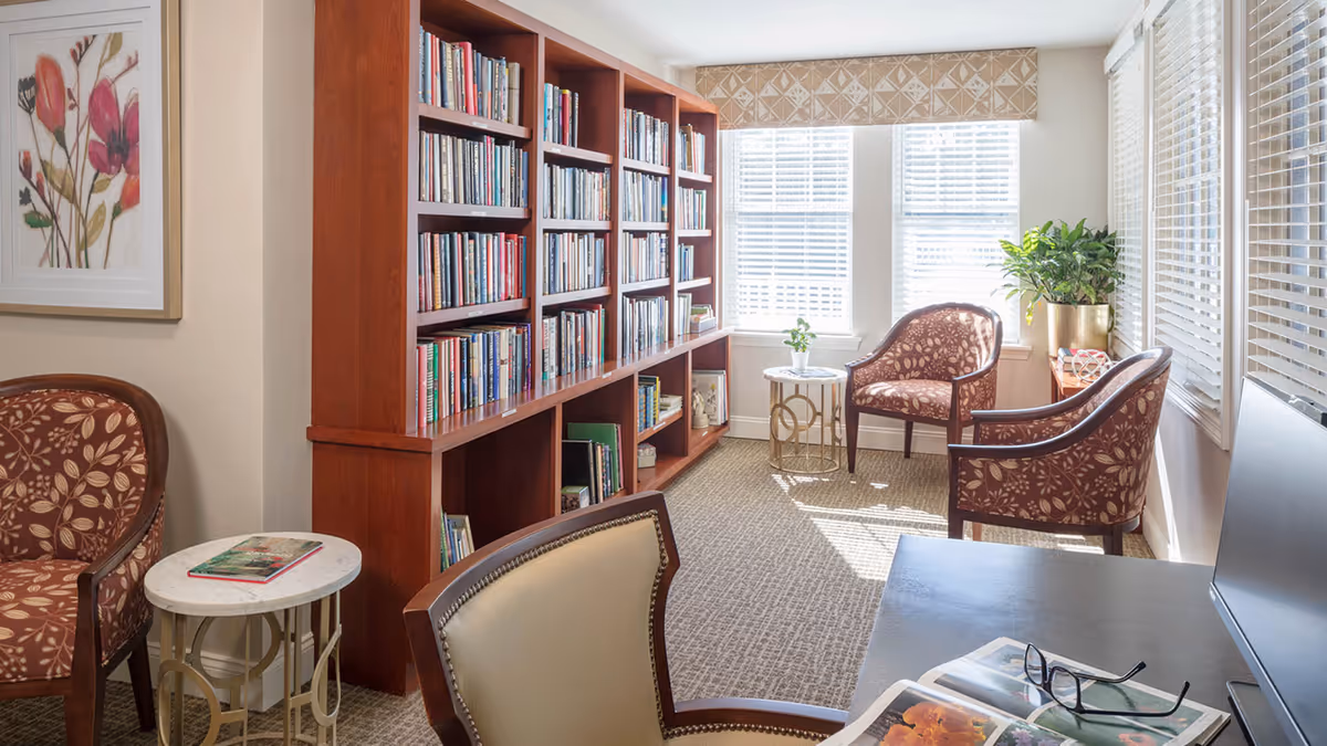 A bright and cozy reading room with a large wooden bookshelf filled with books along one wall. There are three upholstered armchairs with floral patterns arranged around small round tables. Sunlight streams in through multiple windows with white blinds, and a green potted plant sits on a table near the window. A framed floral artwork hangs on the wall, and a desk with reading glasses and an open book is visible in the foreground.