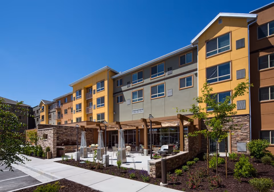 Exterior view of a multi-story senior living facility building with yellow, gray, and brown sections. The building has large windows and a patio area with outdoor seating, pergolas, and tall outdoor heaters. There are landscaped plants and trees around the patio and sidewalk under a clear blue sky.