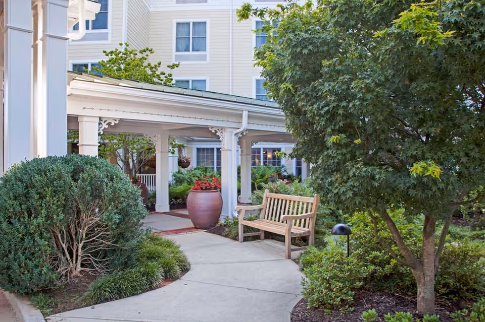Covered building entrance with a wooden bench, large potted planters, and landscaped greenery.