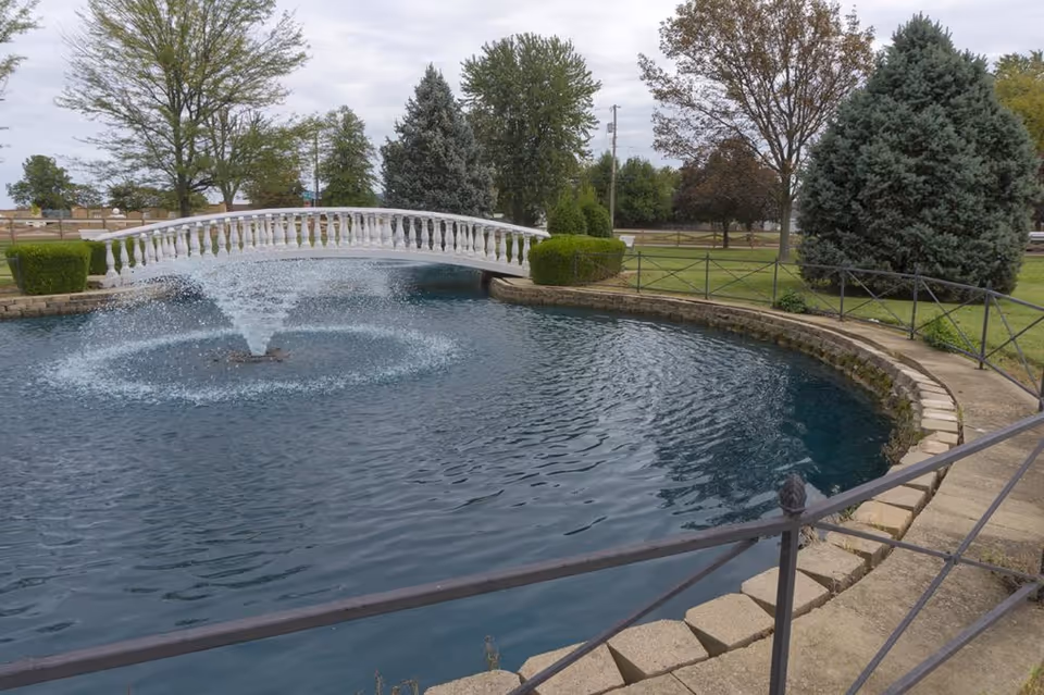 A landscaped outdoor pond with a water fountain spraying water upwards in the center. A white decorative footbridge arches over the pond. Surrounding the pond are trimmed bushes, trees, and a metal fence along a stone walkway.