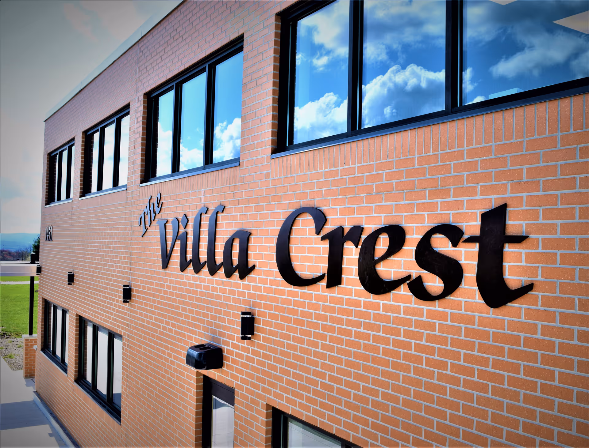 Exterior view of a brick building with large black letters spelling 'The Villa Crest' mounted on the wall under several windows reflecting a blue sky with clouds.