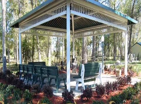 Outdoor covered seating area with green benches arranged under a metal roof structure surrounded by trees and landscaped garden beds with mulch and shrubs.