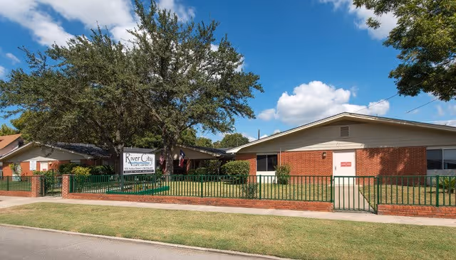 Front exterior of a single-story brick senior care facility with a lawn, fence, sign, and large tree under a blue sky.