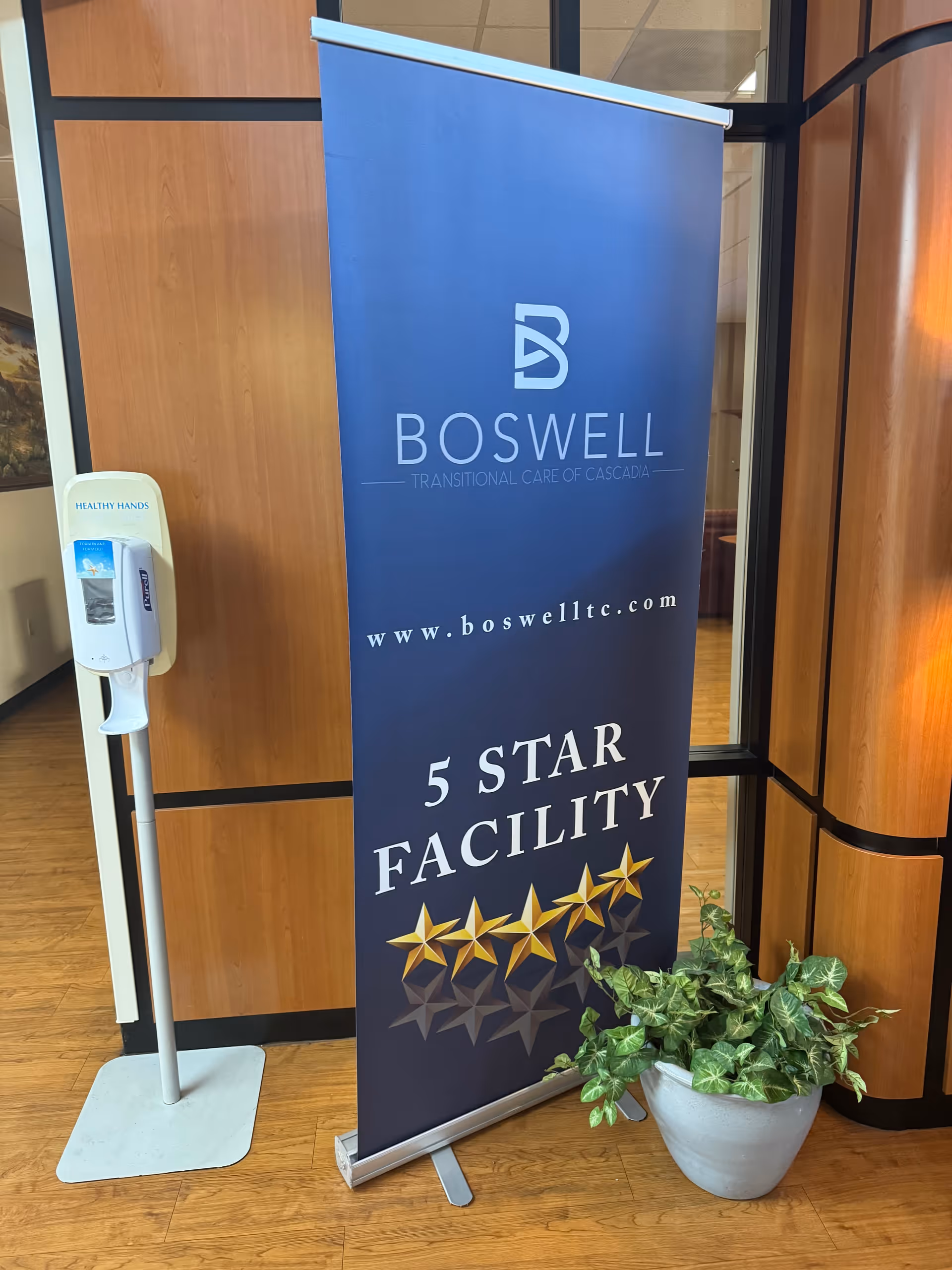 A freestanding blue banner in a facility lobby reading 'Boswell Transitional Care of Cascadia' and '5 Star Facility' with gold stars, next to a hand sanitizer dispenser and a potted plant.