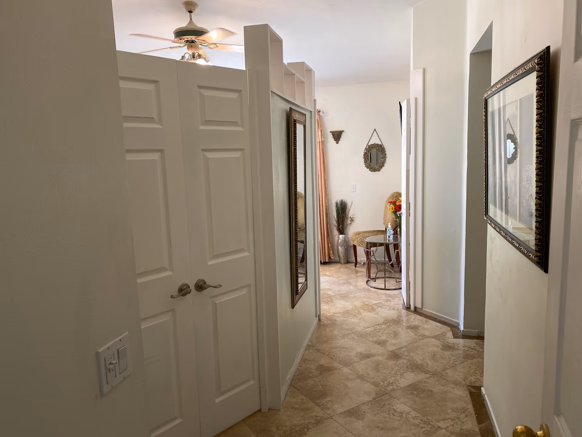 A hallway with beige tiled floor and white walls. On the left side, there are double white doors with silver handles and a light switch on the wall. A large rectangular mirror is mounted on the right wall of the hallway. At the end of the hallway, there is a small seating area with a cushioned chair, a round glass-top table with a bottle of hand sanitizer, a vase with decorative plants, and a small ornate mirror hanging on the wall. A ceiling fan with lights is partially visible above the double doors.