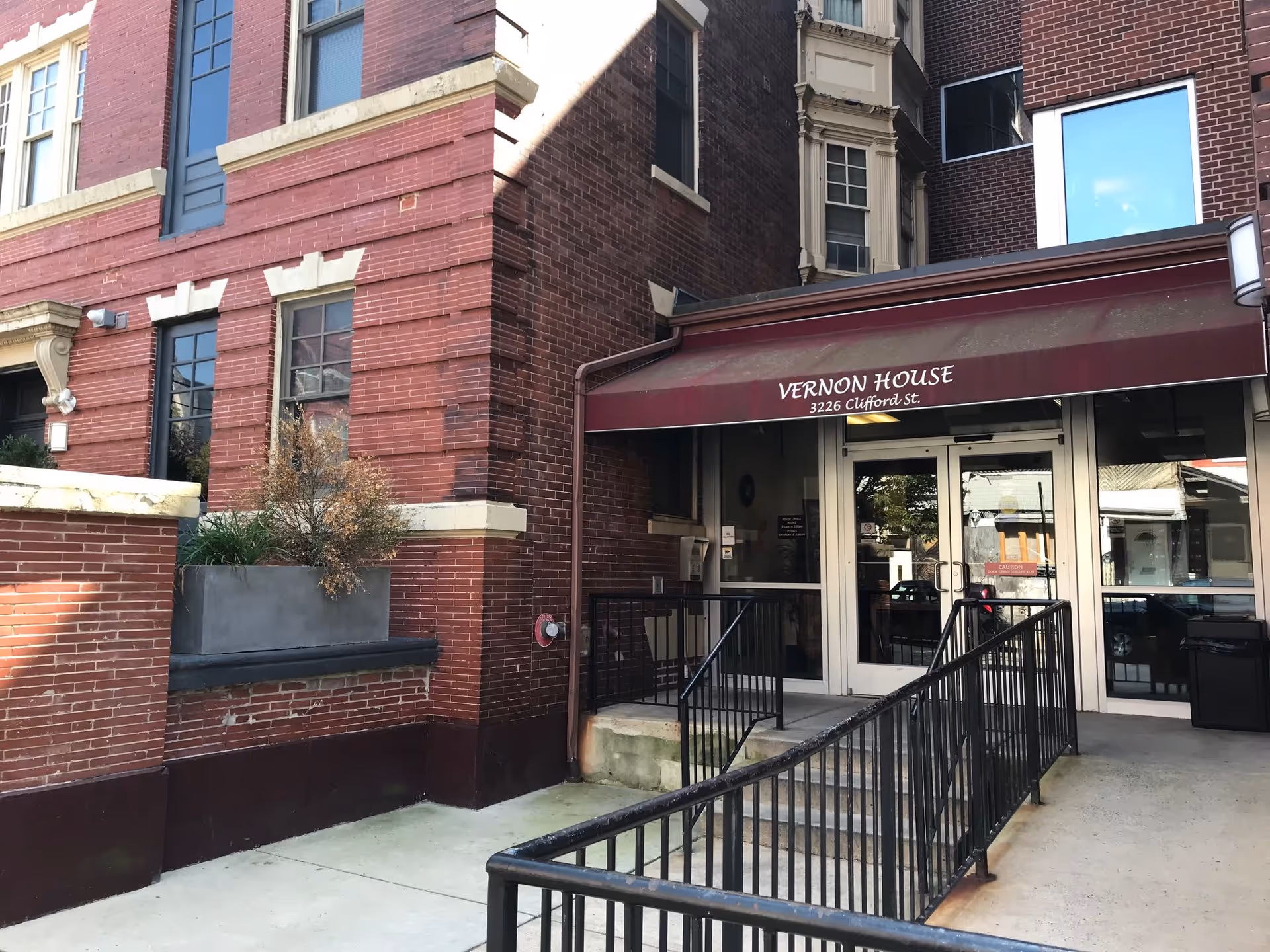 Entrance to Vernon House, a brick building with a maroon awning displaying the name and address 3226 Clifford St. There is a ramp with black railings leading up to glass double doors.