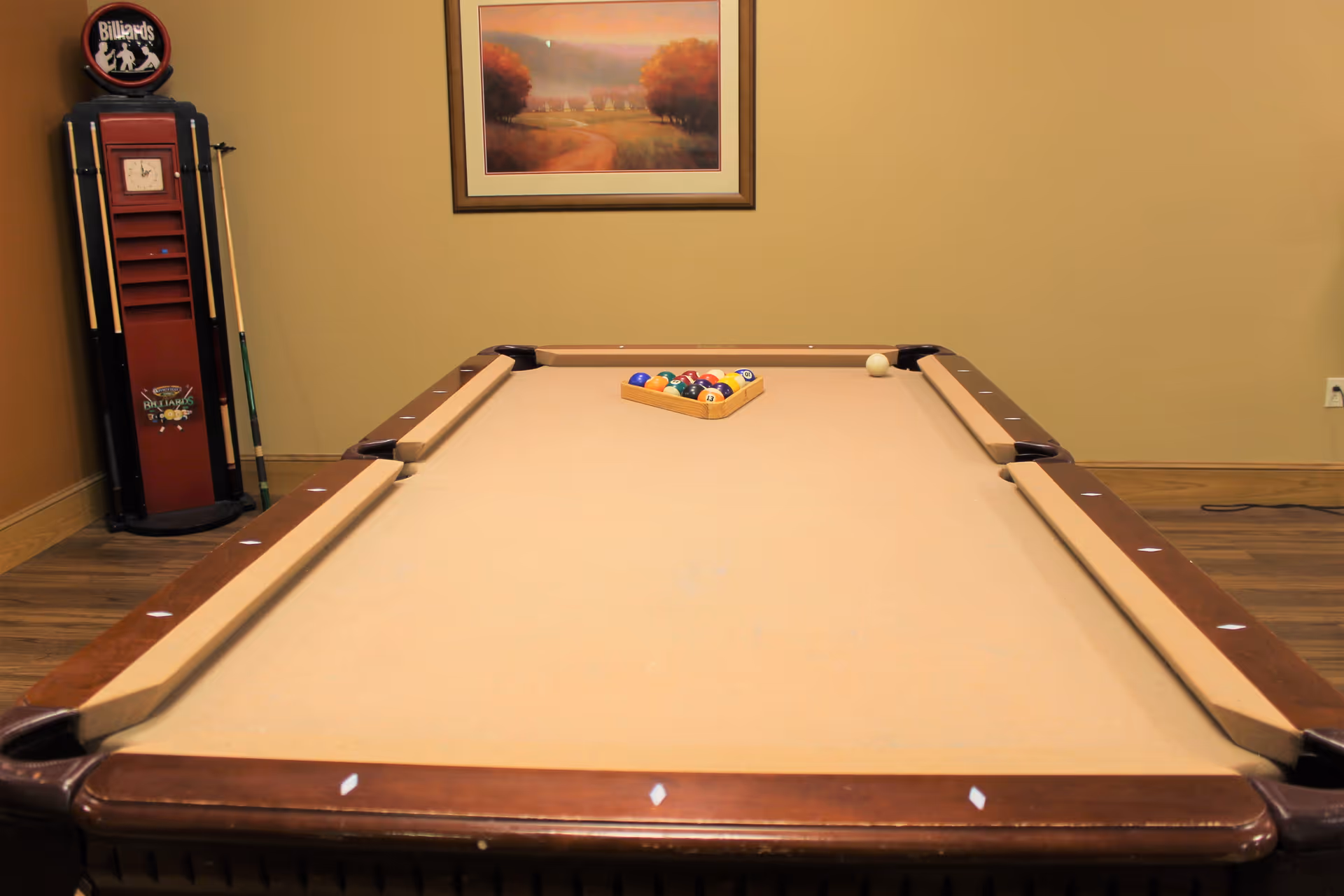 A billiards room with a pool table, racked balls, a cue rack and a framed landscape painting on the wall.