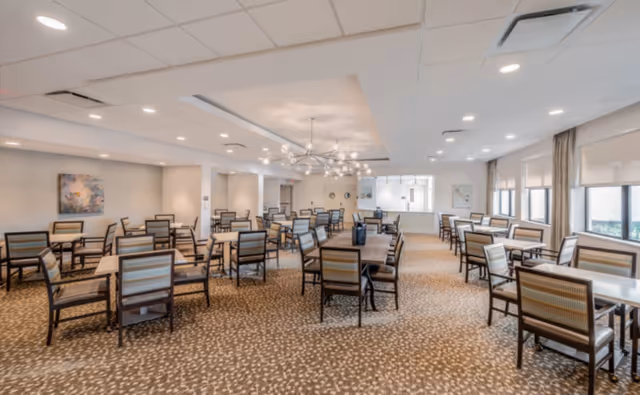Spacious dining room with multiple tables and chairs, patterned carpet, large windows, and modern chandeliers.