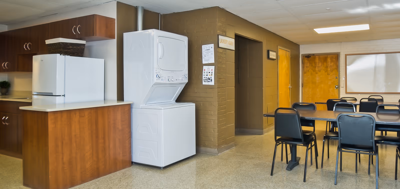 Interior room with a kitchenette area featuring wooden cabinets, a white refrigerator, and a stacked washer and dryer. Adjacent to the kitchenette is a room with tables and black chairs arranged for seating. The walls are painted brown and white, with a door and a whiteboard visible in the background.