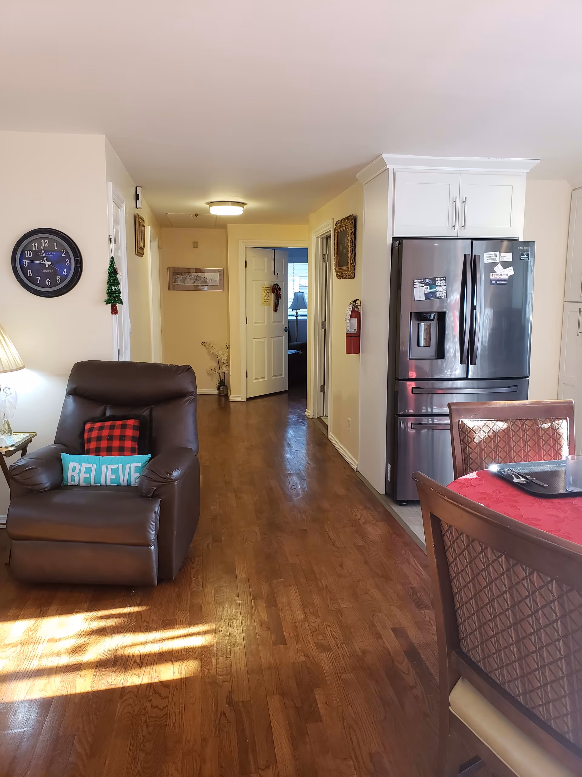 Interior view of a senior living facility showing a hallway with wooden flooring. On the left, there is a brown recliner chair with two pillows, one red and black checkered and one turquoise with the word 'BELIEVE'. A round wall clock is mounted on the wall above the chair. On the right, there is a stainless steel refrigerator next to a dining table with a red tablecloth and wooden chairs. The hallway leads to a room with an open door and a window with blinds.