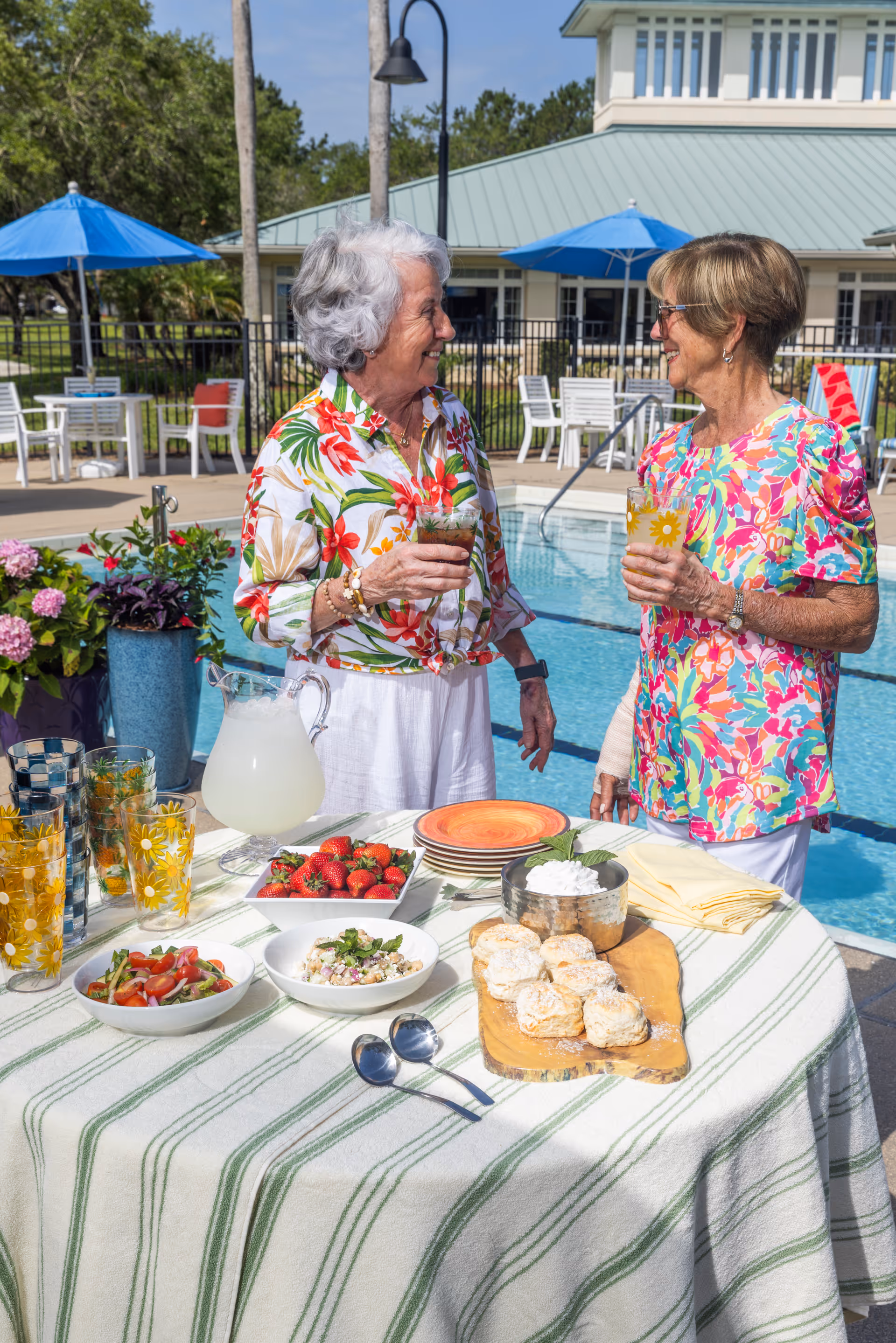 Two elderly women standing and smiling while holding drinks near a poolside table set with plates, glasses, a pitcher of lemonade, strawberries, salad, and biscuits. The background shows a building with a green roof, blue umbrellas, and white chairs around the pool area.