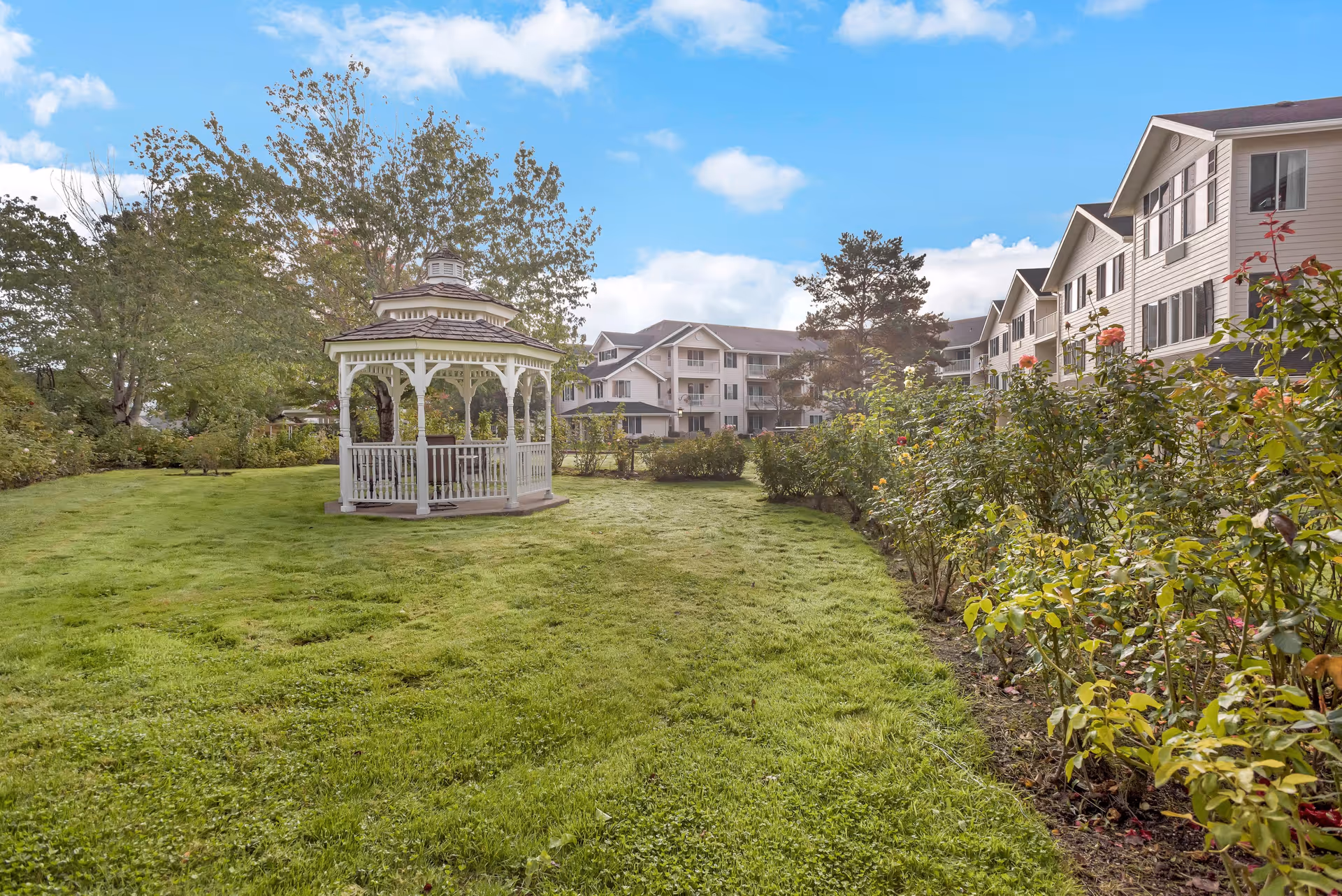 A well-maintained outdoor garden area with a white gazebo on a grassy lawn, surrounded by trees and flowering bushes. In the background, there is a multi-story residential building under a partly cloudy blue sky.