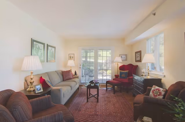 A cozy living room with a beige sofa, two armchairs, a red wingback chair with an ottoman, and a wooden coffee table on a patterned rug. The room is softly lit by table lamps and natural light coming through glass doors and windows. There are framed pictures on the walls and a dark wooden chest of drawers under the window.