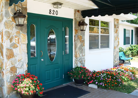 Front entrance of Manatee River Assisted Living with green double doors numbered 820, stone facade, and potted flowers.