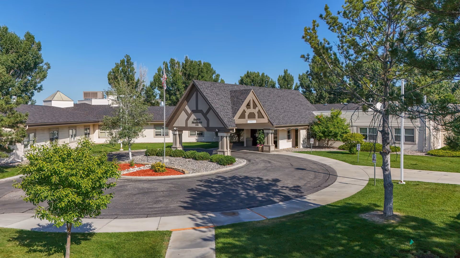 Front exterior of a single-story senior living building with a covered entrance, circular driveway, flagpole, and landscaped lawn.