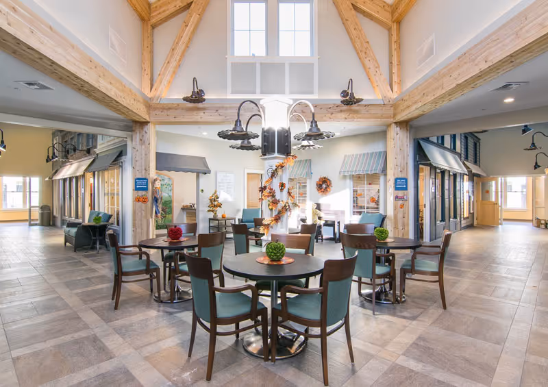 A bright and spacious common area in a senior living facility with round tables and chairs arranged for seating. The room features high vaulted ceilings with exposed wooden beams and large windows letting in natural light. There are decorative plants on the tables and autumn-themed decorations on the central column. The space has a welcoming and comfortable atmosphere with additional seating areas along the walls.