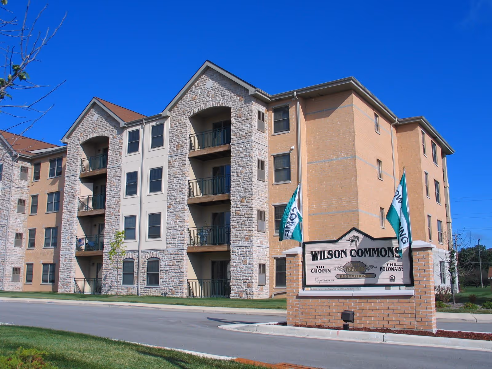 Exterior view of a multi-story senior living facility named Wilson Commons with a brick and stone facade, balconies, and a sign in front displaying the facility's name and flags on either side.