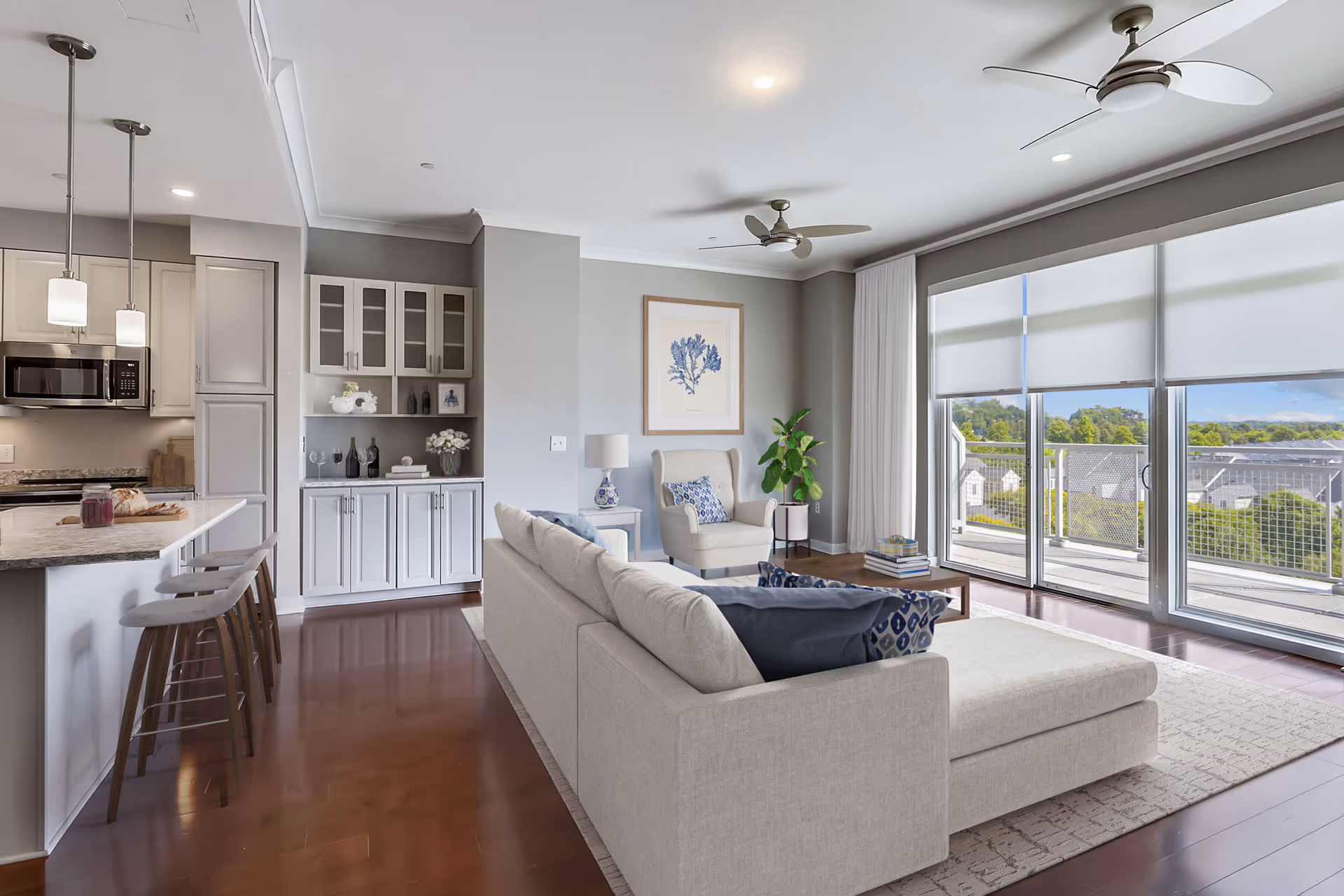 Modern living room and kitchen area with light gray walls, a large beige sectional sofa with blue and patterned pillows, a beige armchair, a coffee table with books, and a large window with roller shades overlooking a balcony and greenery outside. The kitchen features a marble countertop island with three bar stools, pendant lights, white cabinetry, and a microwave.