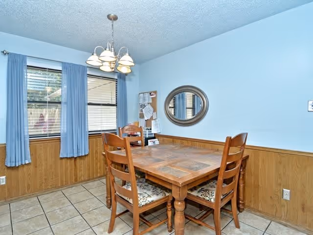 A dining room with a wooden table and four chairs with patterned cushions. The room has light blue walls with wood paneling on the lower half, two windows with blue curtains, a hanging light fixture above the table, a round mirror on the wall, and a bulletin board with papers pinned to it.