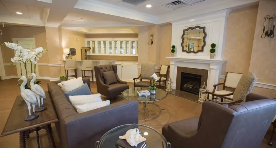 A cozy living room area in a senior living facility with beige walls and a white fireplace. The room features a dark brown leather sofa with white and blue pillows, two matching armchairs, and several wooden chairs with cushions. There are glass-top coffee tables with decorative items, including flowers and seashells. The room is softly lit with wall sconces and recessed ceiling lights, and a decorative mirror hangs above the fireplace.