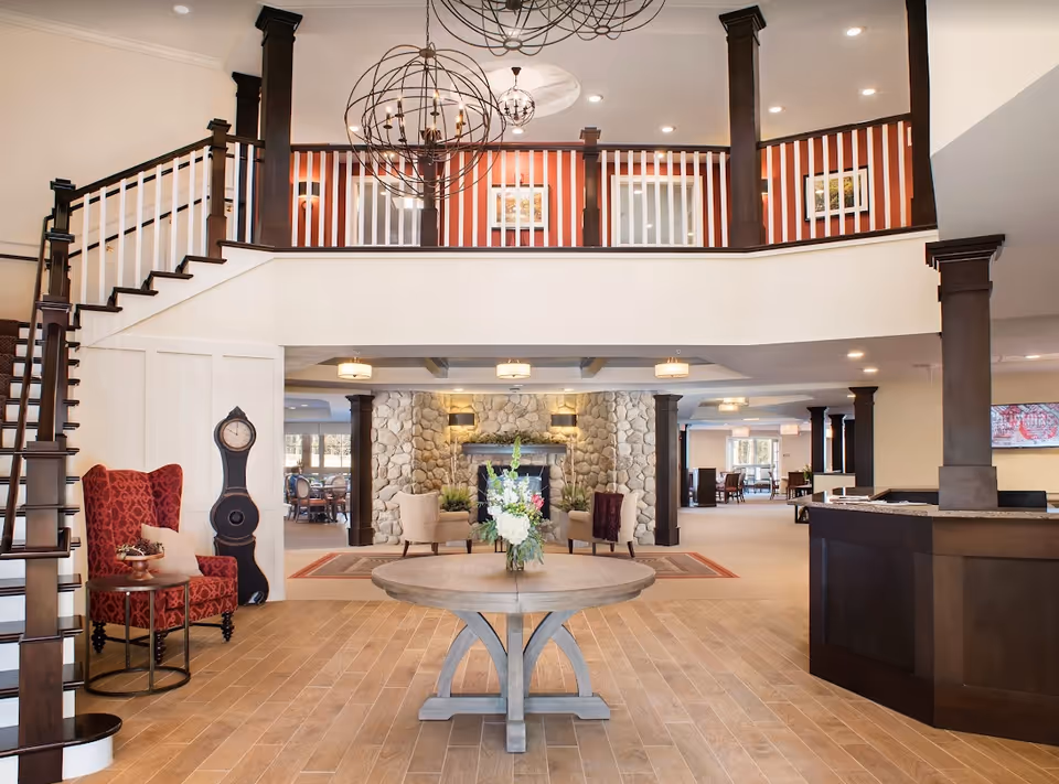Spacious and elegant senior living facility lobby with a round wooden table holding a floral arrangement in the center. To the left, there is a red patterned armchair with a small side table and a tall decorative clock. A staircase with dark wood railing leads to an upper balcony with white spindles and dark wood posts. The back wall features a large stone fireplace flanked by two beige armchairs. The reception desk is on the right side, and the area is well-lit with modern chandeliers and recessed lighting.