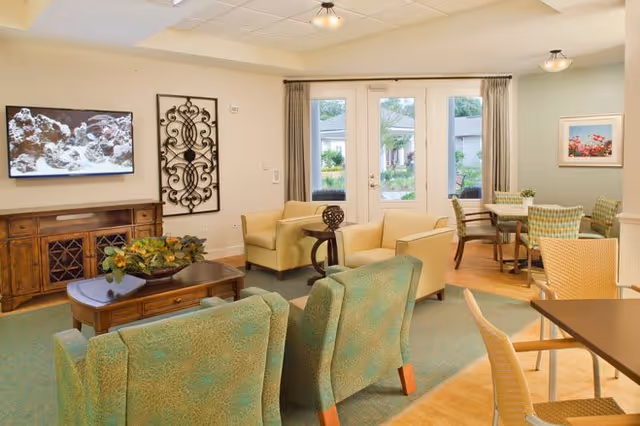 A bright and cozy living room area in Ortega Gardens featuring comfortable seating including green upholstered chairs, beige armchairs, and a wooden coffee table with a decorative plant. A flat-screen TV is mounted on the wall above a wooden cabinet. French doors with curtains open to an outdoor garden area, and a dining table with chairs is visible in the background.
