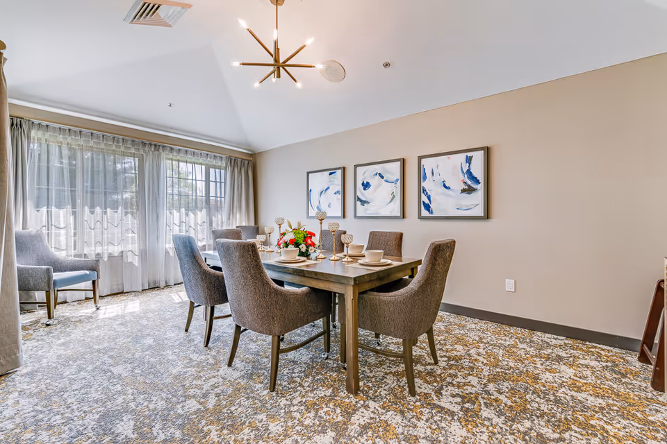 A bright dining room with a rectangular wooden table set for six with beige chairs. The table is decorated with a floral centerpiece and several candle holders. The room has large windows with sheer curtains allowing natural light to fill the space. Three abstract blue and white paintings hang on the beige wall. The floor is covered with a patterned carpet in shades of gray and beige, and a modern chandelier hangs from the ceiling.