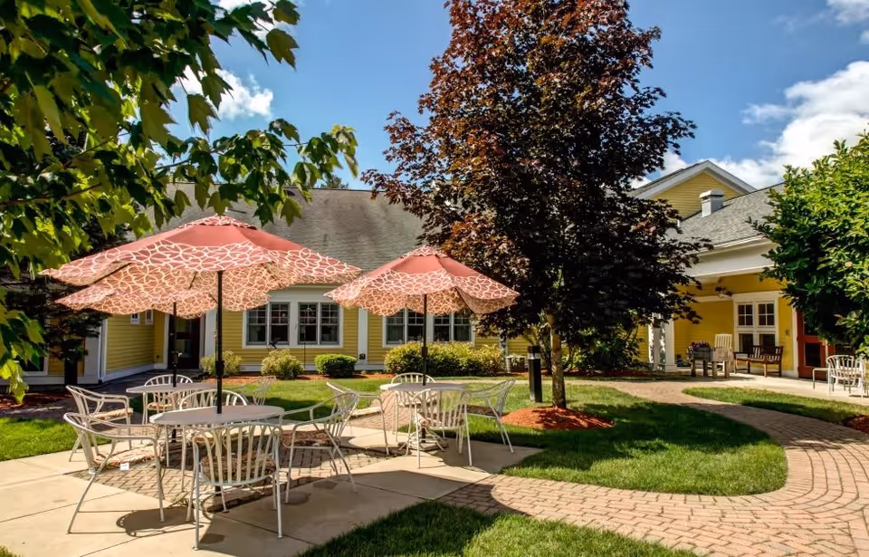 Outdoor patio area at The Arbors of Bedford with round tables and chairs under patterned pink umbrellas, surrounded by green grass, trees, and a yellow building with white trim in the background under a blue sky with some clouds.