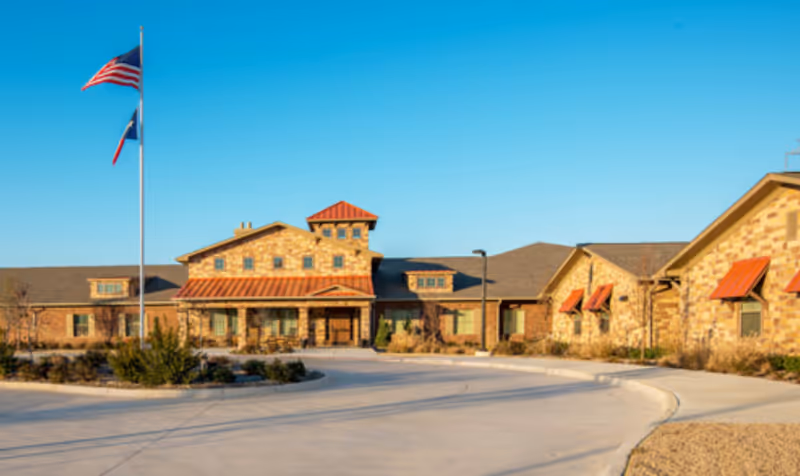 Exterior view of Willow Bend Assisted Living & Memory Care building with stone and brick facade, red awnings, and a circular driveway. Two flagpoles with American and Texas flags are visible in front of the building under a clear blue sky.