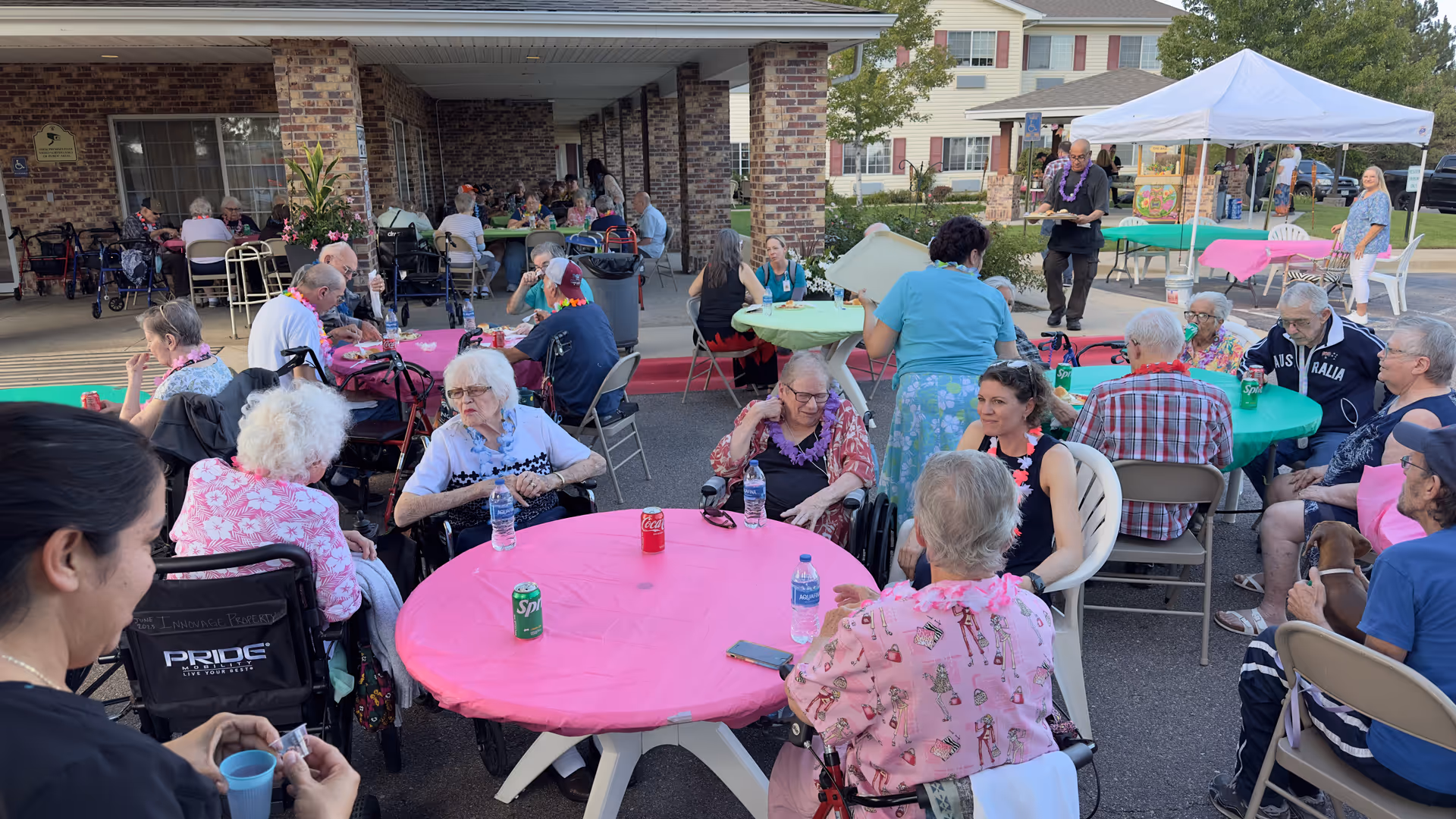 A group of elderly people and caregivers gathered outdoors at Crossroads Senior Living at Northglenn, sitting around tables covered with colorful tablecloths, enjoying drinks and socializing under a covered patio and a white canopy tent.