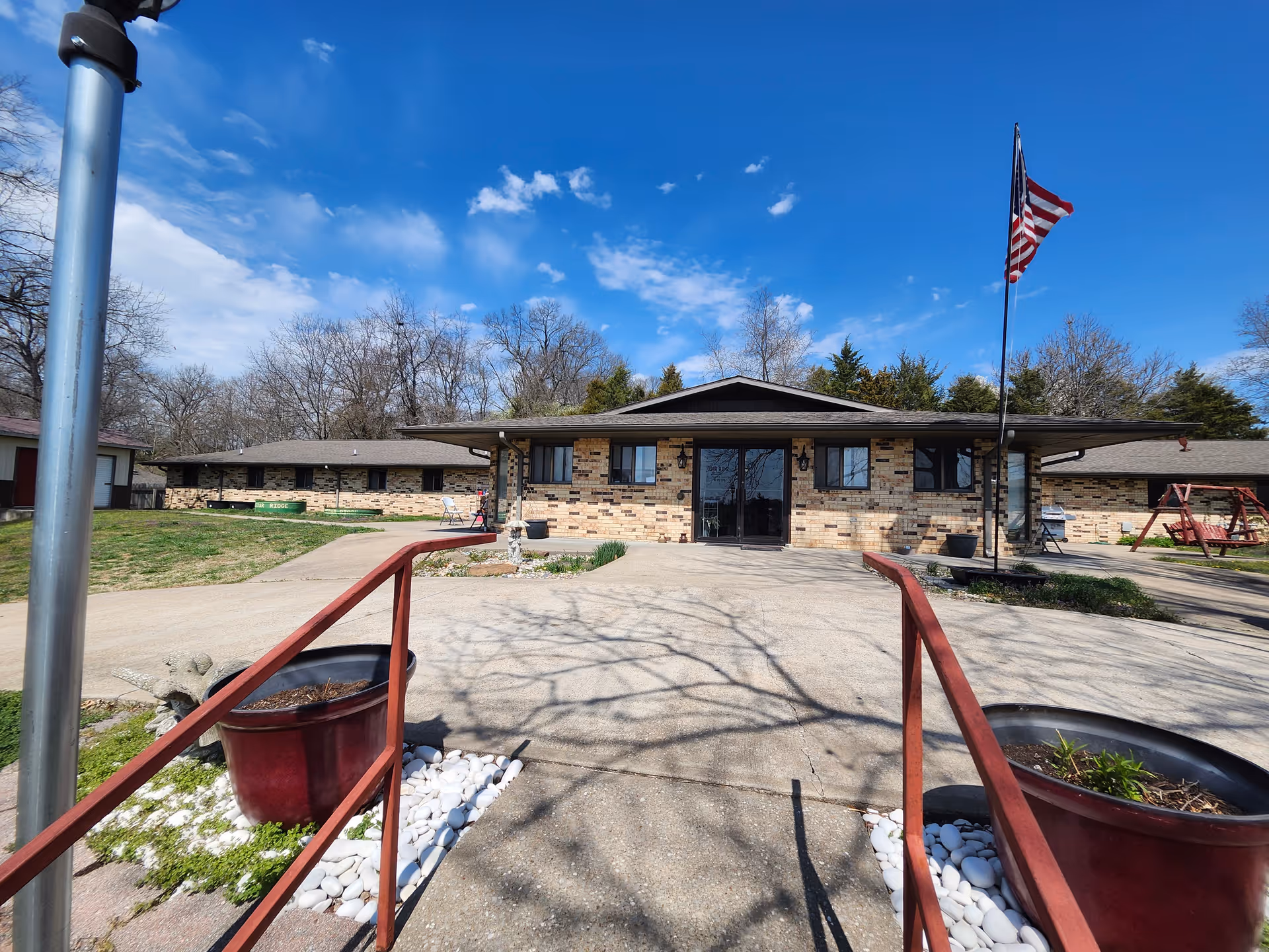 Front entrance of a single-story brick care facility with a walkway, planters, an American flag, and a ramp handrail.