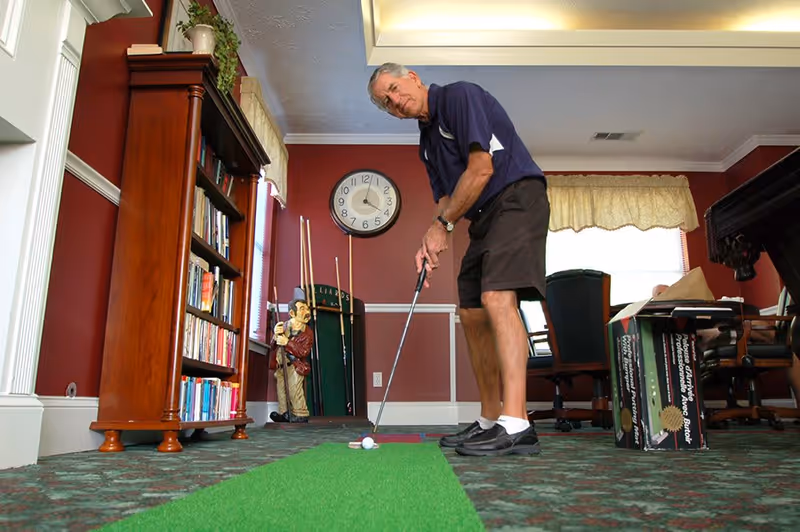 An elderly man playing indoor mini-golf on a green putting mat in a room with red walls, a bookshelf, a clock, and a billiards cue rack. The room has carpeted flooring and a window with a yellow valance.