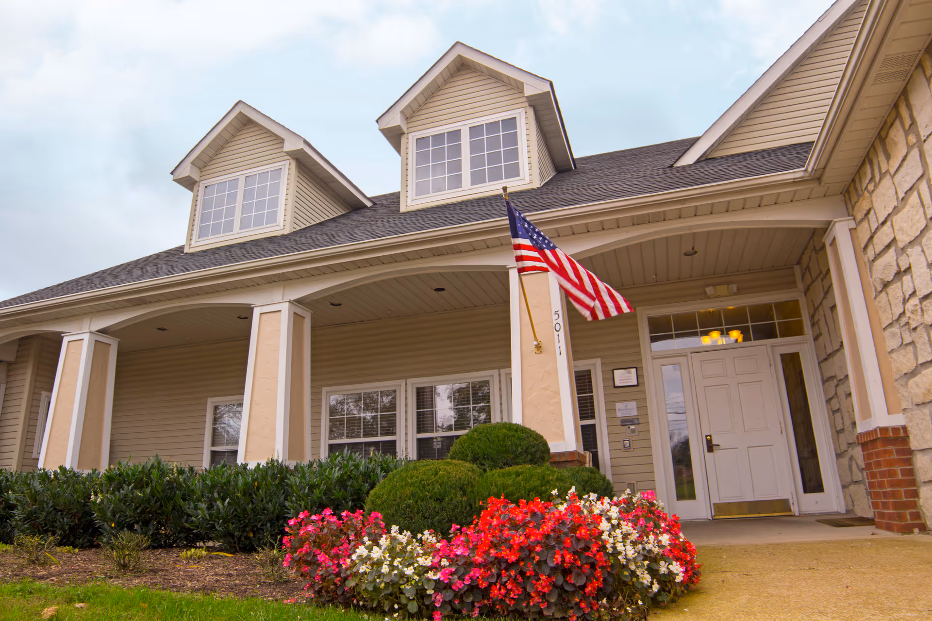 Front exterior view of a building with beige siding, two dormer windows on the roof, an American flag mounted on a column, and a flower bed with red, pink, and white flowers in front of the entrance.