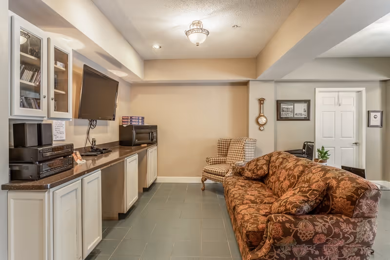 A cozy common living area with a patterned sofa and armchair facing a wall-mounted TV above cabinets and a microwave.