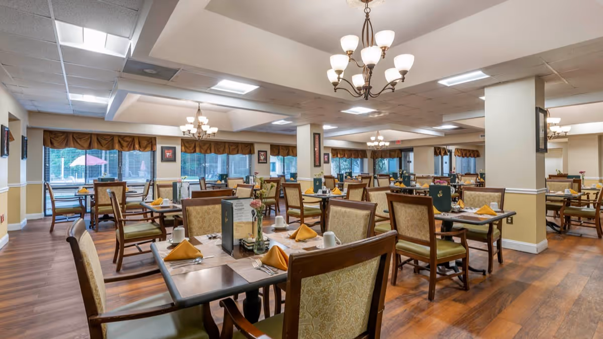 A spacious dining room with multiple tables set for meals, each table featuring yellow folded napkins, white cups, and silverware. The room has wooden flooring, beige walls with white trim, large windows with brown valances, and chandeliers hanging from the ceiling providing warm lighting.
