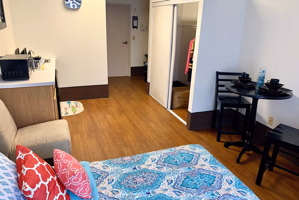 Interior view of a supportive living facility room with a bed covered in a blue and white patterned bedspread and red pillows, a small beige sofa, a kitchenette area with a microwave, and a small round dining table with two chairs and black dishware. The room has wood flooring and a closet with sliding doors.