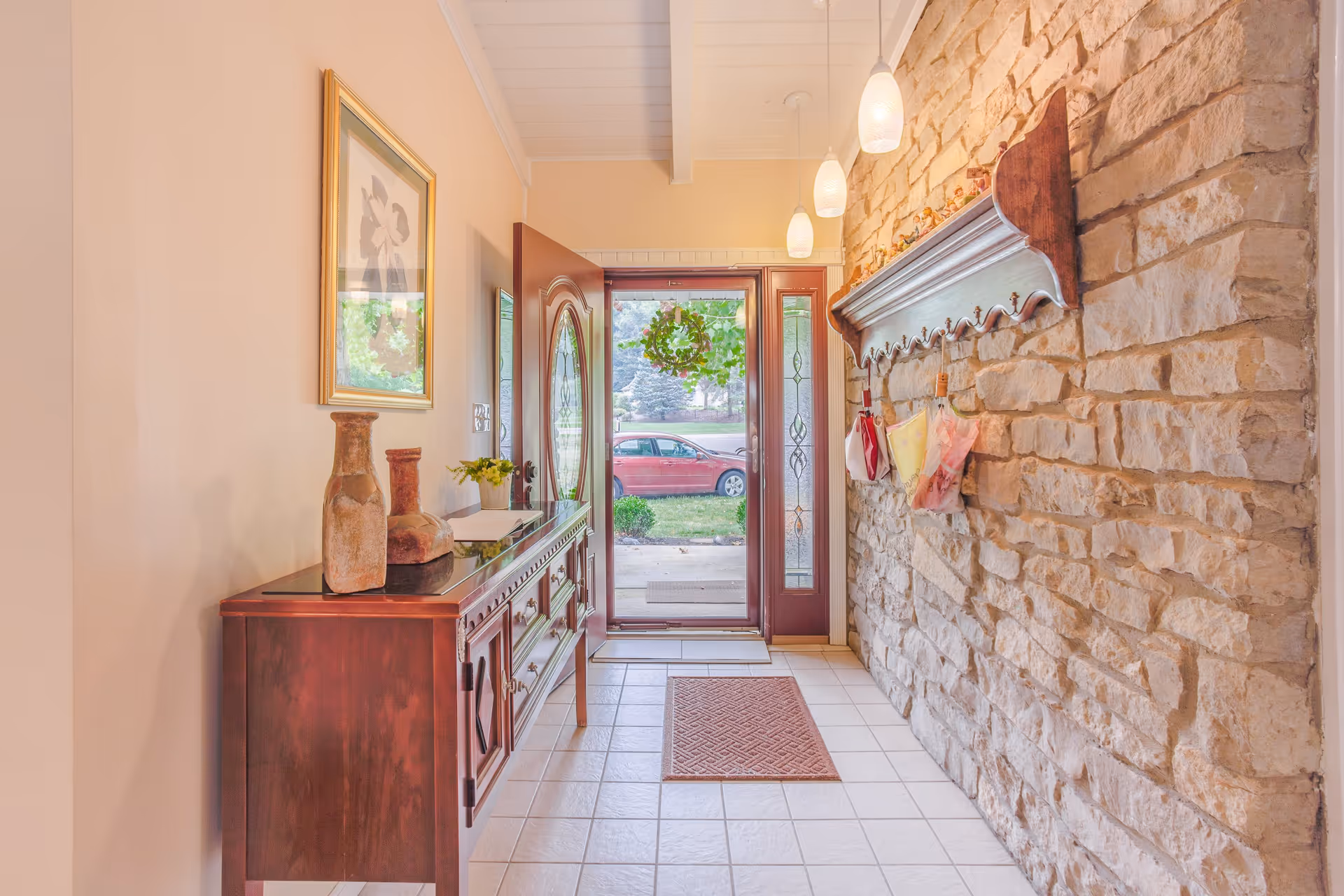 A bright entryway with a glass-paneled front door and sidelights, a wooden console table with decorative vases and a small plant, a stone accent wall with a wooden shelf holding small items and hanging bags, and three pendant lights hanging from the ceiling.