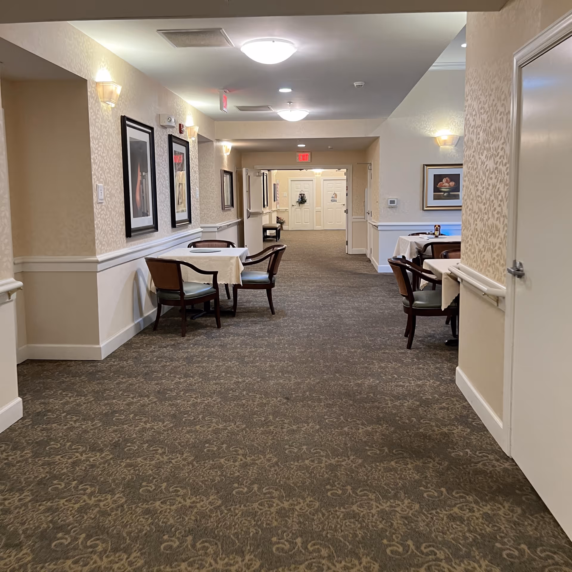 A carpeted hallway in an assisted living facility with tables and chairs set along the walls. The walls are decorated with framed artwork and wall sconces providing soft lighting. The hallway leads to a set of double doors at the far end.