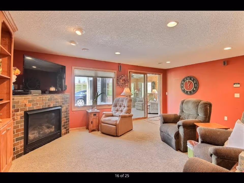 A cozy living room with red walls, beige carpet, and recessed ceiling lights. The room features a flat-screen TV mounted above a brick fireplace, a wooden side table with a lamp, and three comfortable armchairs arranged around the space. There is a large window and sliding glass door that provide a view of the outdoors.