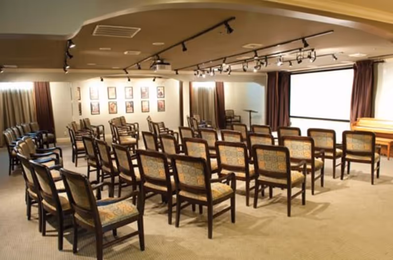 A conference or presentation room with multiple rows of wooden chairs with patterned cushions facing a large white projection screen. The room has beige walls, carpeted floor, framed pictures on one wall, and ceiling track lighting.