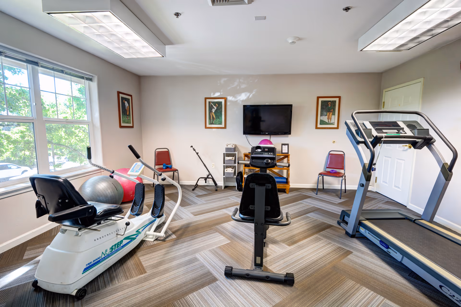 A small fitness room with exercise equipment including a recumbent bike, a treadmill, and an elliptical machine. The room has large windows letting in natural light, three framed pictures on the wall, a flat-screen TV mounted on the wall, and two red chairs placed against the walls. The floor is carpeted with a patterned design.