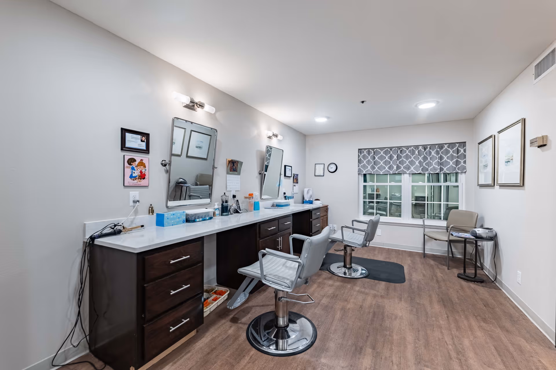 Interior view of a senior living facility's hair salon area with two styling chairs in front of a long counter with mirrors, hair care products, and tissue boxes. The room has wood flooring, a window with patterned valance, and a small seating area with two chairs and a side table.