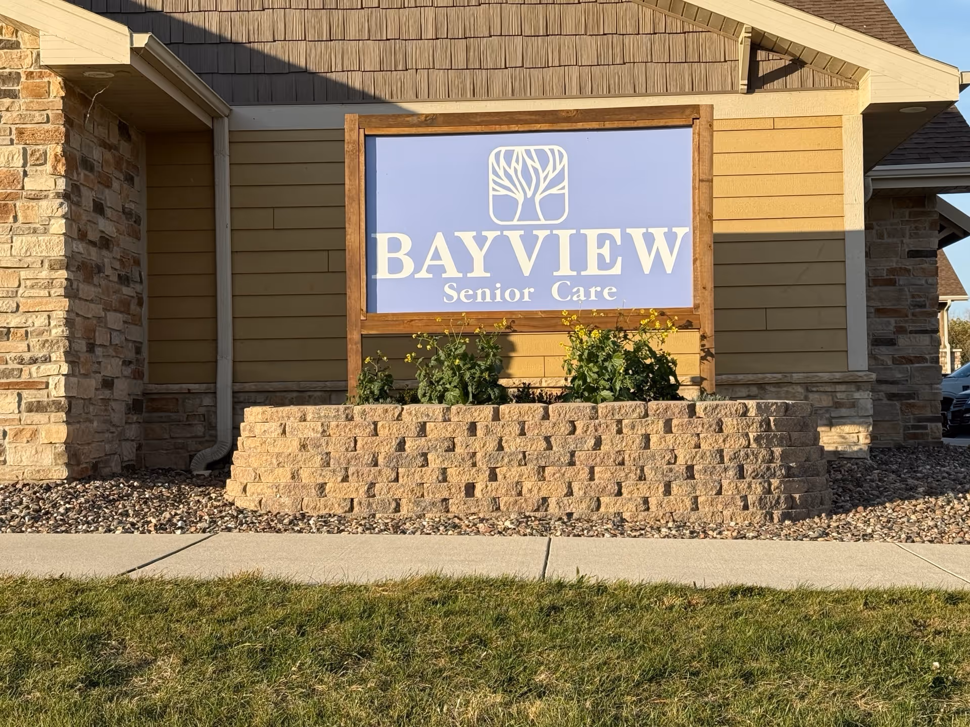 Outdoor view of a Bayview Senior Care sign mounted on a wooden frame in front of a building with stone and yellow siding. The sign features a tree logo above the text. There is a small garden bed with plants and flowers in front of the sign, bordered by a low stone wall. A sidewalk and grass lawn are visible in the foreground.