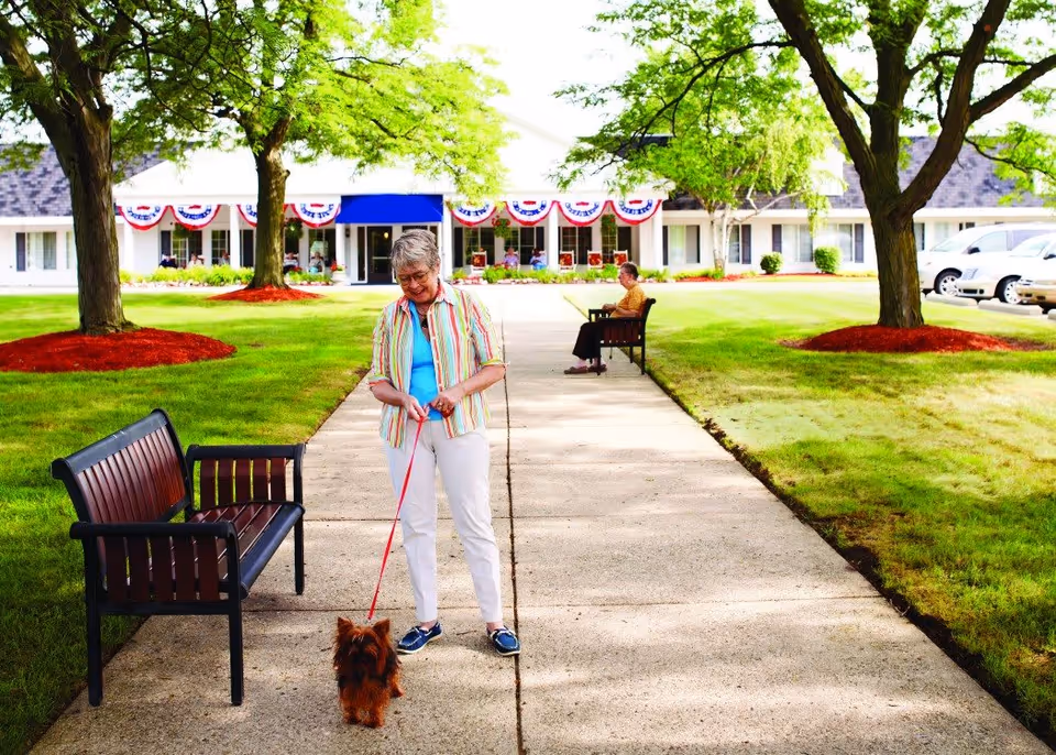 An elderly woman walking a small dog on a leash along a paved pathway in a green outdoor area with trees and benches. Another elderly person is sitting on a bench in the background near a building decorated with red, white, and blue bunting.